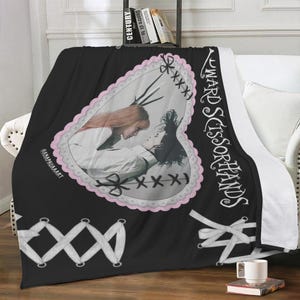 Puede incluir: Manta negra con un diseño en forma de corazón que presenta una imagen en escala de grises de dos personas. La manta tiene el texto "Edward Scissorhands" en blanco, junto con costuras decorativas y lazos. La manta está sobre un sofá blanco.