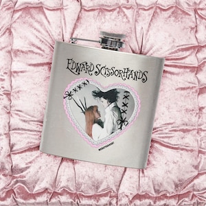 Puede incluir: Una petaca de acero inoxidable plateado con el texto "Edward Scissorhands" y una imagen en forma de corazón de los personajes de la película. El corazón tiene un borde rosa y lazos negros. La petaca tiene una tapa plateada, sobre un fondo rosa texturizado.