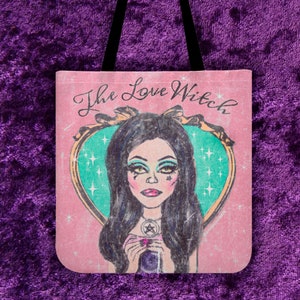 May include: A pink tote bag with a vintage illustration of a woman with dark hair and a heart-shaped mirror. The text "The Love Witch" is written in pink and white lettering.