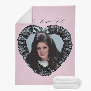 May include: A pink blanket featuring a portrait of a woman within a heart-shaped frame, adorned with black lace and a white bow. The text "'s Iconic Doll" is printed at the top. A folded white blanket is at the bottom.