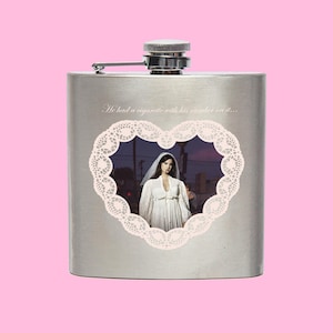 Puede incluir: Una petaca de acero inoxidable con tapa plateada y cierre de bisagra. La petaca presenta un diseño en forma de corazón con una mujer con un vestido blanco y el texto "He had a cigarette with his number on it..."