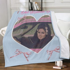 May include: A light blue throw blanket featuring a heart-shaped image of a woman in a car. The image is framed in pink, with pink bows at the corners. The blanket has the text "Hel Mountain" in a script font.
