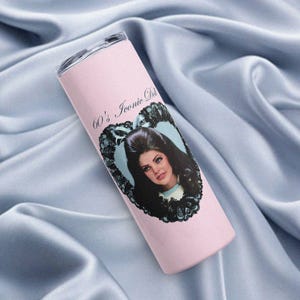 Puede incluir: Un vaso rosa con un retrato de una mujer con cabello oscuro en un marco en forma de corazón con bordes de encaje negro. El texto "60's Iconic Doll" está impreso en el vaso. El vaso tiene una tapa transparente.