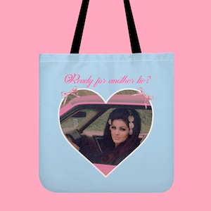 Könnte beinhalten: Hellblaue Tragetasche mit einem herzförmigen Bild einer Frau in einem rosa Auto. Das Bild ist mit einem weißen Rand versehen und der Text "Ready for another lie?" ist in rosa Schrift über dem Herzen geschrieben. Die Tasche hat schwarze Griffe.