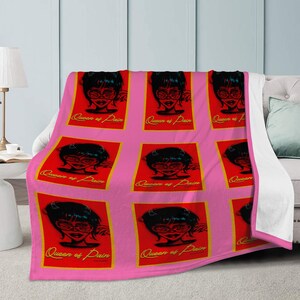 Puede incluir: Manta rosa con un patrón repetido de la cara de una mujer con gafas y el texto "Queen of Pain" sobre un fondo rojo con un borde dorado. La manta está sobre un sofá.