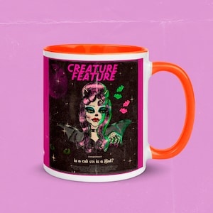 Puede incluir: Una taza de cerámica blanca con un borde y un asa naranja. La taza presenta una ilustración de estilo retro de una mujer con alas de murciélago y el texto "CREATURE FEATURE is a cat or is a bat?"