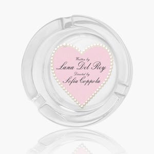 May include: A clear glass ashtray with a pink heart design in the centre. The heart is outlined with white pearls and features the text "Written by Lana Del Rey, Directed by Sofia Coppola."