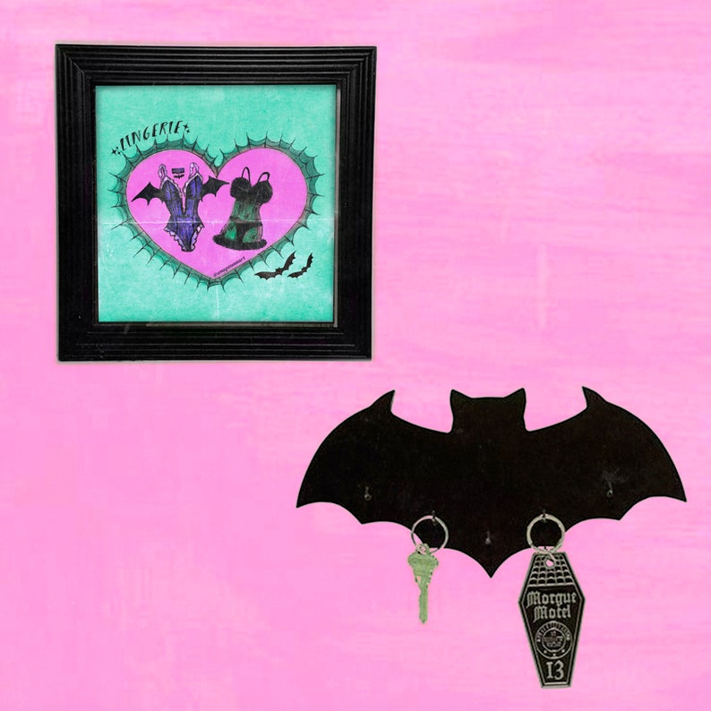 May include: A black bat-shaped key holder with three hooks. The key holder is mounted on a pink wall. There are two keys hanging from the hooks, one is a silver key and the other is a silver key with a coffin-shaped tag that says "Morgue Motel 13". A framed picture with a heart and lingerie design is hanging on the wall above the key holder.