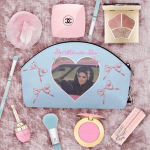 Pode incluir: Uma coleção de produtos de beleza, incluindo uma bolsa de cosméticos azul claro com uma imagem em forma de coração de uma mulher em um carro, blush rosa, sombra para os olhos e um compacto Chanel rosa. Os itens estão dispostos em uma superfície rosa texturizada.