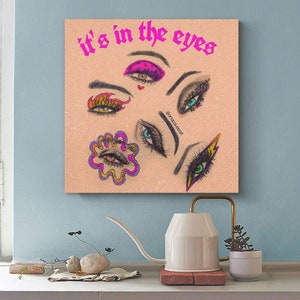 May include: A pink and white canvas print with the text "it's in the eyes" in black. The print features a collage of colorful eyes with different makeup styles.