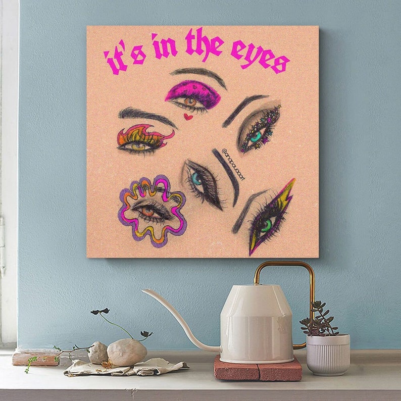 May include: A pink and white canvas print with the text "it's in the eyes" in black. The print features a collage of colorful eyes with different makeup styles.