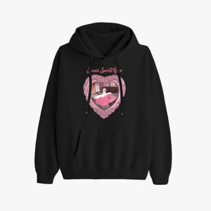 Puede incluir: Sudadera con capucha negra con bolsillo delantero y cordón. Presenta un gráfico en forma de corazón rosa con el texto "Sweet Serial Killer" y una ilustración de una persona en una cama.