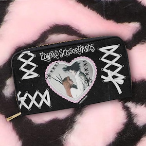 Puede incluir: Cartera negra con cremallera con el texto "Edward Scissorhands". Una imagen en forma de corazón de los personajes de la película está centrada en la cartera. Costuras blancas en forma de cruz acentúan el diseño. Una cremallera y tirador dorados completan el look.