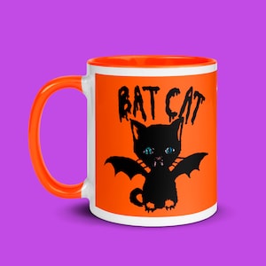Puede incluir: Taza de cerámica naranja y blanca con una ilustración de un gato murciélago negro y el texto "BAT CAT" en la parte delantera.