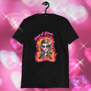 Op de afbeelding: Zwart T-shirt met een afbeelding van een vrouw met lang blond haar in vlammen. De tekst "Fire of Glam" staat boven het hoofd van de vrouw.