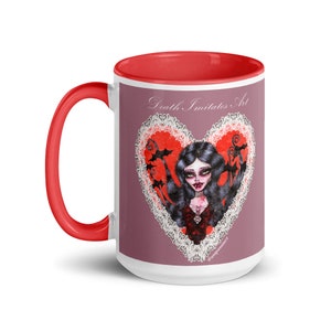 Peut inclure: Mug en céramique blanche avec intérieur et poignée rouges. Le mug présente un motif représentant une femme aux longs cheveux noirs et aux lèvres rouges dans un cœur rouge avec une bordure en dentelle blanche. Le texte "Death Imitates Art" est imprimé au-dessus du cœur.