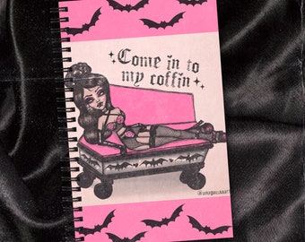Puede incluir: Cuaderno rosa con una ilustración en blanco y negro de una mujer tumbada en un ataúd con el texto "Come in to my coffin". El fondo es rosa con murciélagos negros.