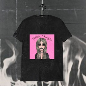 May include: Black t-shirt with a pink square graphic featuring a drawing of a woman with long blonde hair and the text "TAYLOR MOMSEN" in black.