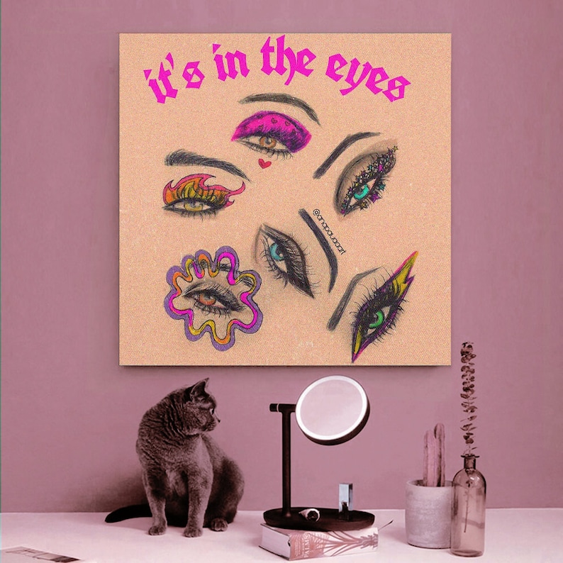 May include: A beige canvas print with the text "it's in the eyes" in pink letters. The print features a collage of colorful eyes with different makeup styles.