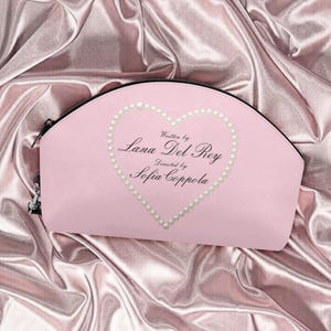 May include: A pale pink cosmetic bag with a black zip and a heart-shaped pearl design. The text "Written by Lana Del Rey, Directed by Sofia Coppola" is inside the heart. The bag rests on a pink satin surface.