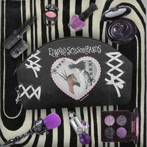 Puede incluir: Bolsa de maquillaje negra con el texto "Edward Scissorhands" y una imagen en forma de corazón de los personajes de la película. La bolsa está rodeada de varios artículos de maquillaje, incluyendo lápiz labial, sombra de ojos, esmalte de uñas y un pincel.