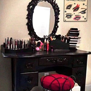 May include: A black vanity table with a large ornate mirror and a red cushioned stool. The table has three drawers and is decorated with intricate carvings. The mirror has a white oval frame with a decorative design. The vanity table is set up with various makeup and beauty products.
