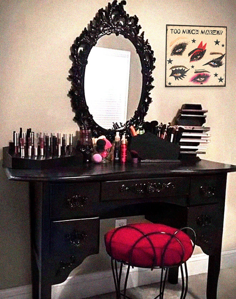 May include: A black vanity table with a large ornate mirror and a red cushioned stool. The table has three drawers and is decorated with intricate carvings. The mirror has a white oval frame with a decorative design. The vanity table is set up with various makeup and beauty products.