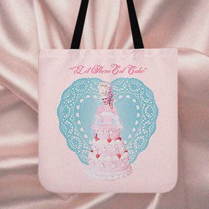 Può includere: Borsa tote rosa con un'illustrazione a forma di torta di una donna in abito storico, incorniciata da un centrino blu a forma di cuore. Il testo "Let Them Eat Cake" è stampato sopra l'immagine. La borsa ha manici neri.