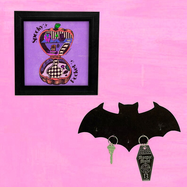 May include: A black bat-shaped key holder with two keys hanging from it. One key is a traditional house key, and the other is a coffin-shaped key with the words "Morgue Motel" and "13" on it. A framed print with a purple background and the words "Spooky Pocket" is hanging on the wall above the key holder.