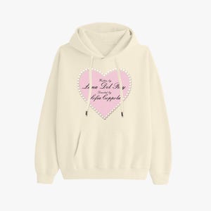 Può includere: Felpa con cappuccio color crema con tasca frontale e cappuccio con coulisse. Una grafica a cuore rosa con finiture perlata e la scritta "Written by Lana Del Rey, Directed by Sofia Coppola" è centrata sul petto.