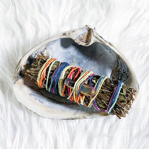 May include: A smudge stick wrapped in colorful string, resting in a white seashell. The smudge stick is made of dried herbs and is used for cleansing and purification.