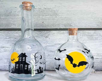 May include: Two glass bottles with cork tops decorated with Halloween themes. One bottle has a black silhouette of a house and a tree with bats flying around a yellow moon. The other bottle has a yellow moon with a black bat and bats flying around the moon.