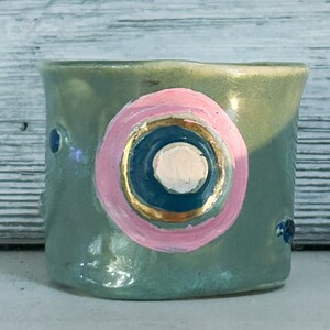 May include: A small, green ceramic cup with a pink, blue, and gold painted design. The design features a circle with a smaller circle inside, and a gold ring around the outer circle.