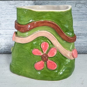 May include: A green ceramic vase with a wavy, multi-colored design. The vase has a pink flower with a brown center.