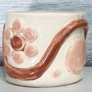 May include: A white ceramic planter with a brown swirl design and pink raised dots forming a flower shape.