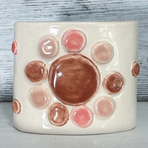 May include: White ceramic vase with a raised design of pink and brown circles. The circles are arranged in a circular pattern.