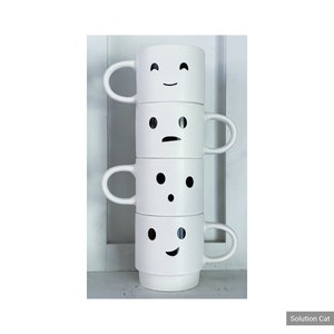 May include: A stack of four white ceramic mugs with different black and white emoji faces. The mugs are stacked on top of each other, with the top mug showing a happy face, the second mug showing a sad face, the third mug showing a surprised face, and the bottom mug showing a smiling face.