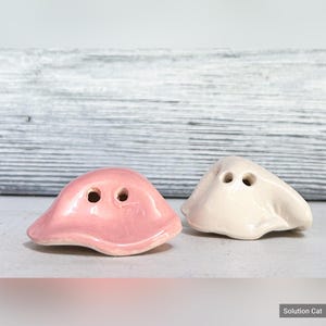 May include: Two ceramic ghost salt and pepper shakers. The ghost on the left is pink and the ghost on the right is white.