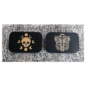May include: Two black rectangular boxes with gold designs. The first box has a gold skull with a crescent moon design. The second box has a gold butterfly skeleton design.