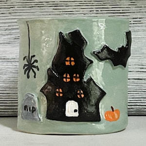 May include: A ceramic mug with a green glaze and a Halloween theme. The mug features a black silhouette of a haunted house with orange windows, a bat, a spider, and a tombstone that says "RIP".