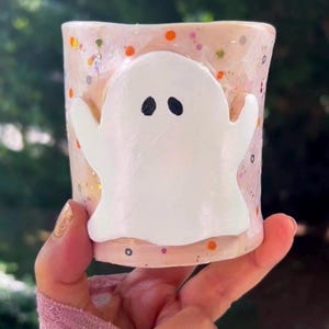 May include: A pink ceramic cup with a white ghost design and orange and white glitter accents.