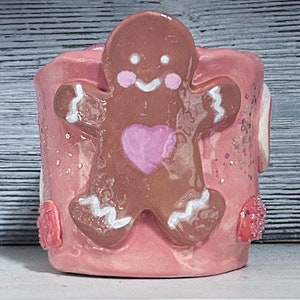 May include: A pink ceramic mug with a gingerbread man design. The gingerbread man is brown with white icing and a pink heart. The mug has a sparkly pink glaze.