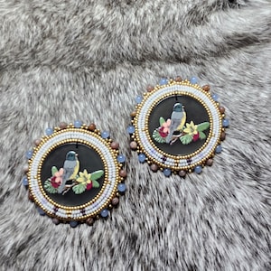 Puede incluir: Dos pendientes redondos con cuentas, con un fondo negro. Cada pendiente presenta un pájaro pintado sobre una rama floral. Los pendientes están enmarcados con cuentas blancas, doradas y azules, sobre un fondo de piel gris.