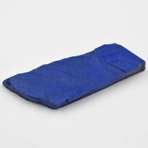 May include: A rectangular piece of deep blue lapis lazuli stone with rough edges. The stone has a slightly textured surface and is displayed against a white background. The stone is approximately 4 inches long.