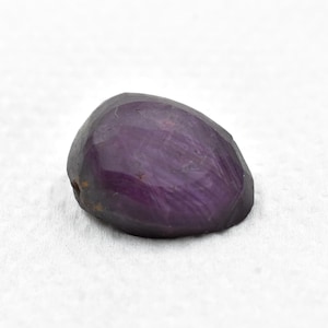 May include: A faceted, deep purple gemstone with an irregular, rounded shape. The stone features a polished, reflective surface with visible internal inclusions and natural variations in color intensity across its translucent, dark violet body.
