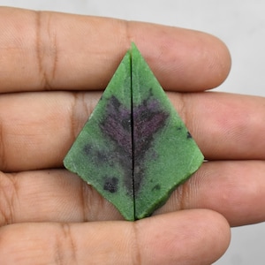 Pode incluir: Duas peças de pedra preciosa de zoisite em bruto, cada uma em forma de losango. As pedras são principalmente verdes com inclusões roxas escuras e pretas. As pedras preciosas são seguradas em uma mão contra um fundo simples.