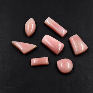 Puede incluir: Una colección de gemas rosas pulidas en varias formas, incluyendo ovaladas, rectangulares y triangulares. Las gemas se exhiben sobre un fondo oscuro, resaltando sus superficies lisas y brillantes y sus sutiles variaciones de color.