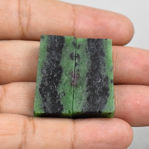 Pode incluir: Duas peças retangulares de pedra preciosa de rubi zoisite. As pedras têm uma cor de base verde com inclusões pretas e vermelho escuro. As pedras preciosas são seguradas em uma mão contra um fundo neutro. As pedras são cortadas e polidas.