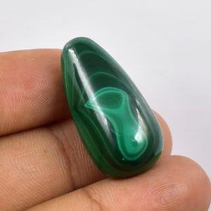 Può includere: Una pietra di malachite lucidata a forma di goccia. La pietra mostra un colore verde intenso con motivi a vortice e bande di verde più chiaro. La superficie è liscia e riflettente, mettendo in risalto la bellezza naturale del minerale.