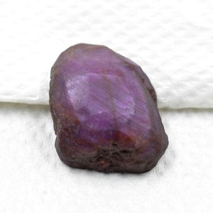 Può includere: Una pietra preziosa grezza e non tagliata con una tonalità viola e magenta profonda. La pietra presenta una superficie superiore sfaccettata e lucida che transita in una base ruvida, strutturata e marrone scuro, mostrando variazioni minerali.